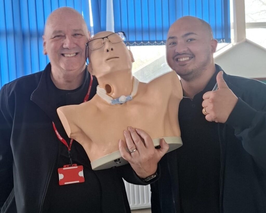 Two men smiling and giving a thumbs up with a CPR manikin