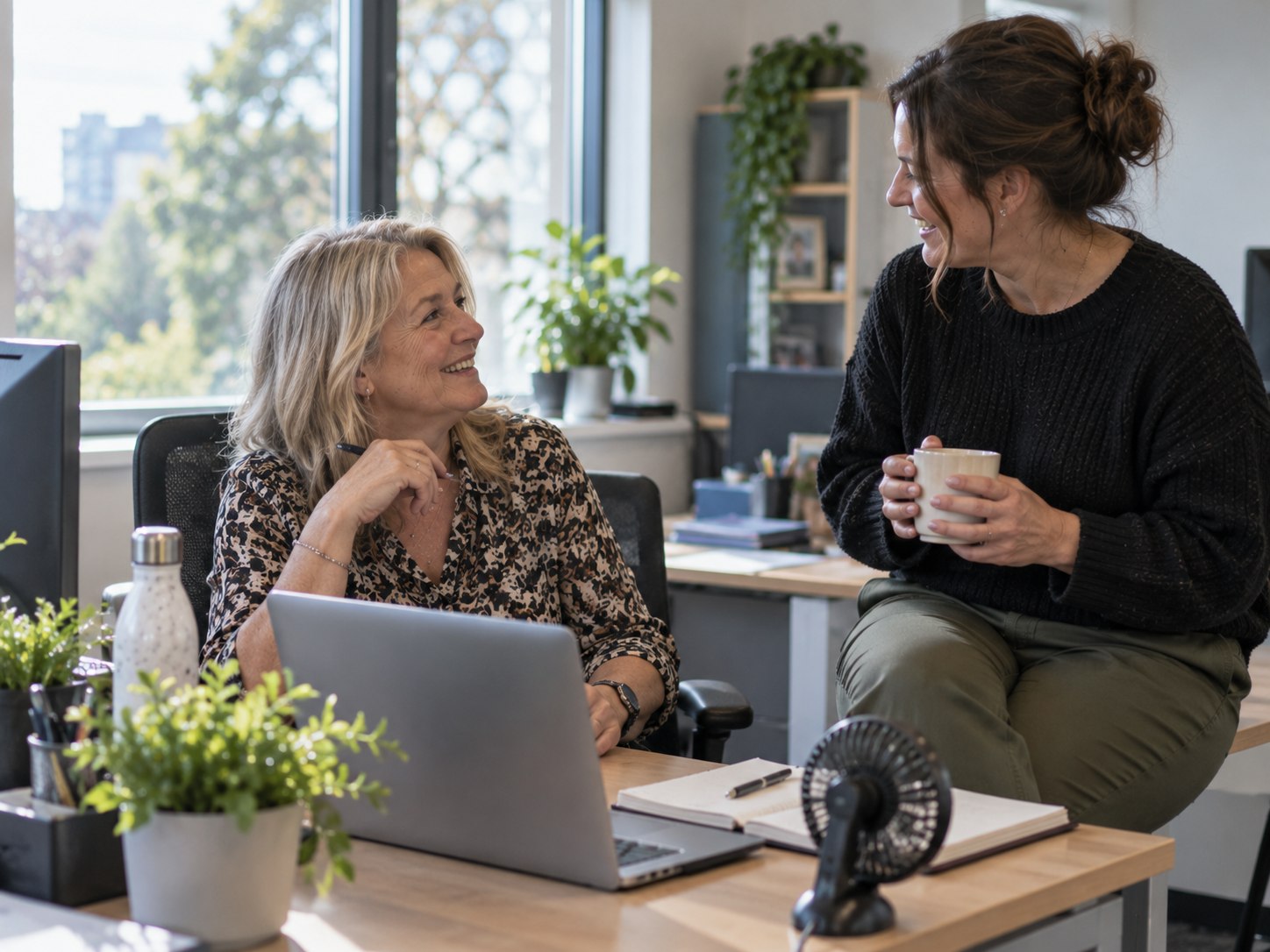 Two colleagues laughing together at a desk in a bright office, one with a mug of tea