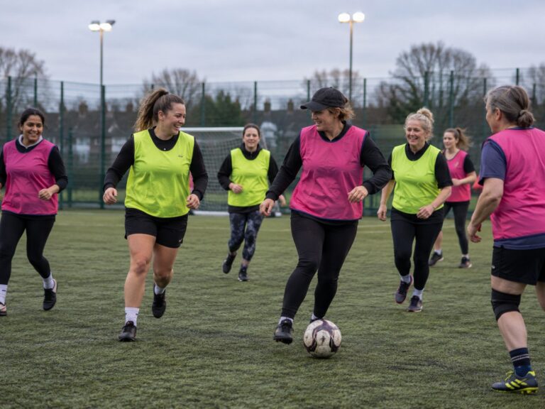 A group of women of different ages playing recreational football together on an astroturf pitch, laughing as they go